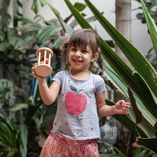 A smiling young girl playing with a handcrafted wooden rattle outdoors, demonstrating the tactile sensory play recommended by pediatricians over screen time.